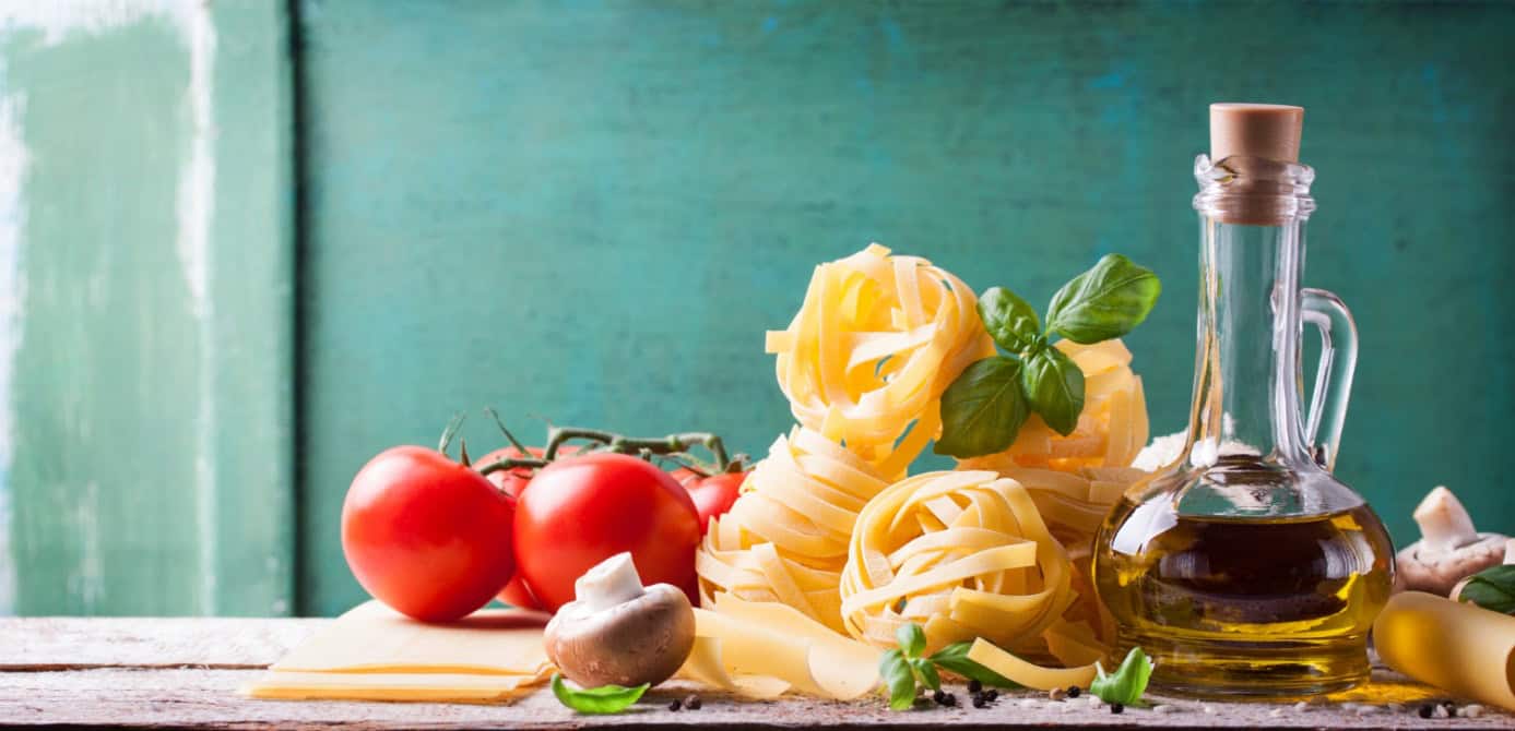 Na stole leży surowy makaron tagliatelle, oliwa, pomidory, pieczarki i świeże zioła